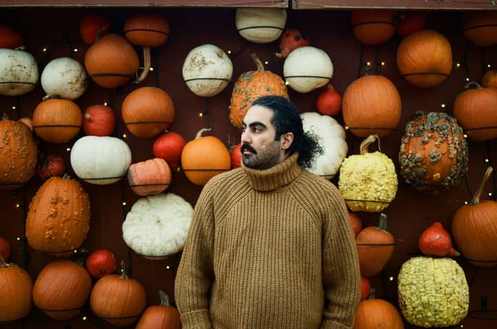Man in a warm sweater stands against a colorful pumpkin wall, showcasing autumn vibes.