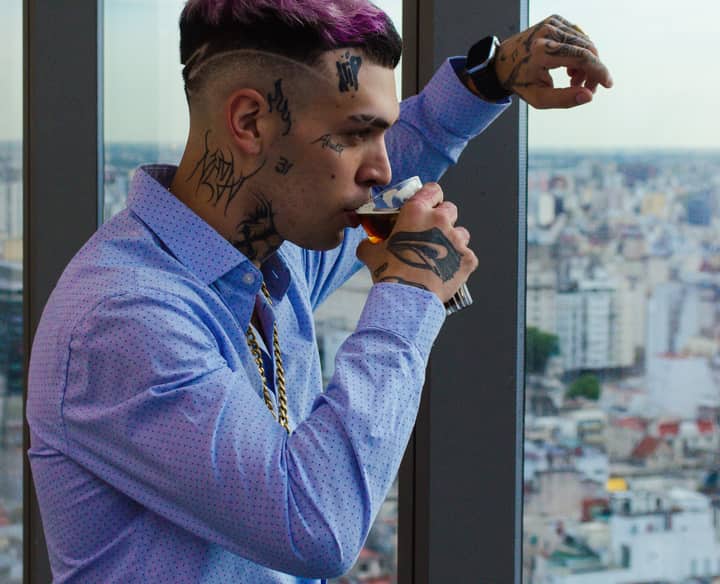 Tattooed man in blue shirt drinking beverage against cityscape backdrop, showcasing urban lifestyle.