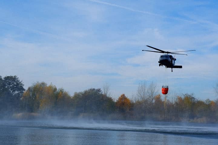 A helicopter gathers water from a lake for firefighting efforts. Blue sky and autumn trees.