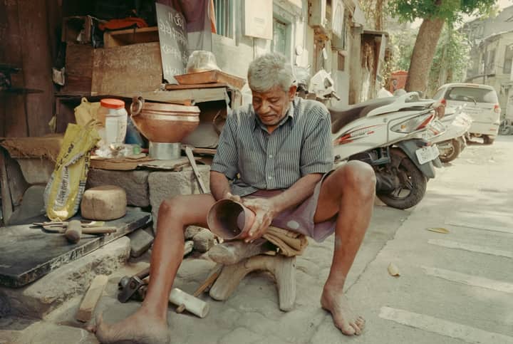 An elderly man crafts pottery on an Indian street, surrounded by tools and motorcycles, during the day.