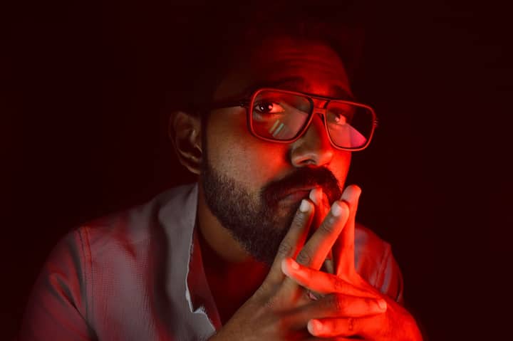 A thoughtful man with red ambient lighting, wearing glasses and a beard, looking upwards.