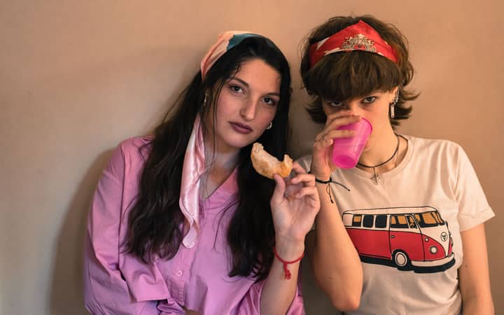 Two stylish young women indoors with a snack and pink drink. Casual and vibrant atmosphere.