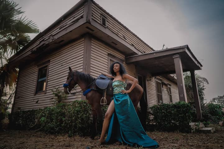 A woman in a glamorous dress poses confidently with a horse beside a rustic wooden house.