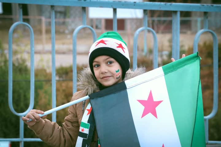 A young girl in Syria proudly holds the Syrian flag, symbolizing hope and resilience.