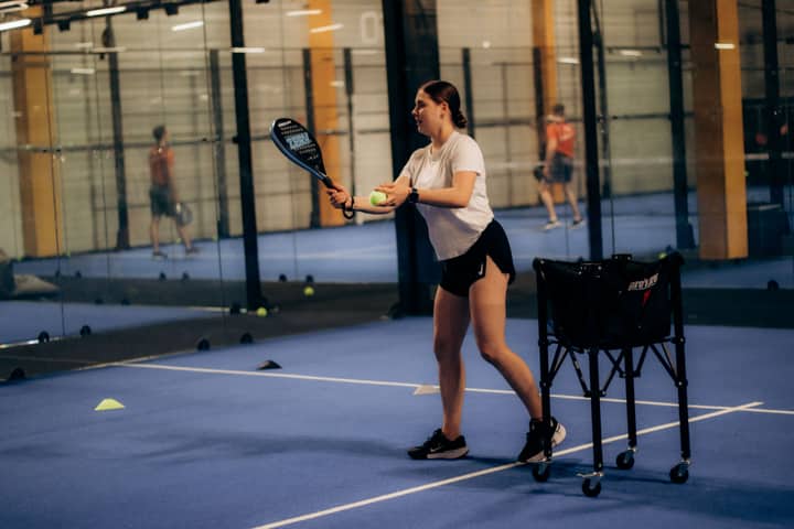 A young woman holding a paddle and ball on an indoor paddle tennis court, ready to serve.