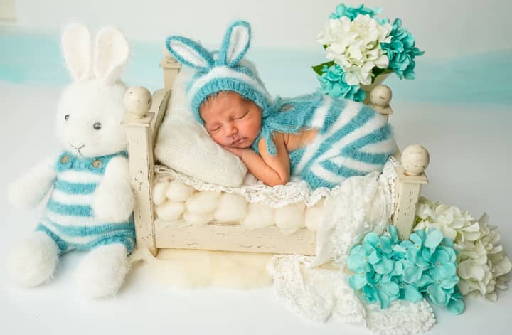 Newborn baby wearing a blue bunny costume asleep on a bed with flowers and a stuffed toy.