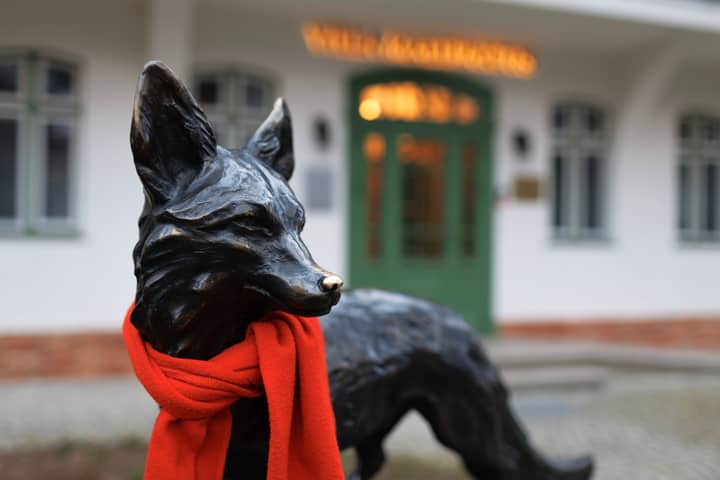 Bronze fox statue adorned with a vibrant red scarf in front of a classic building facade.