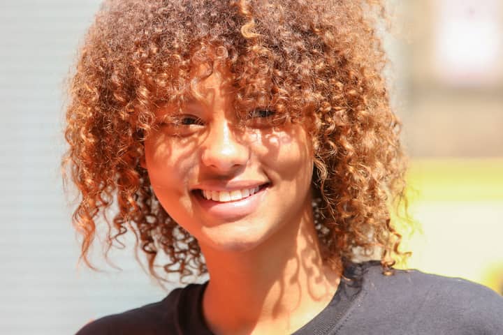 A young woman with curly hair smiles brightly during a sunny day outdoors, radiating joy and warmth.