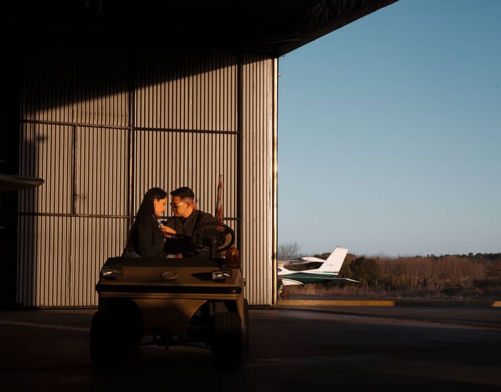 A couple sits inside a hangar, enjoying a moment with an airplane visible outside.