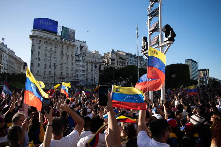 A large crowd waves Venezuelan flags during a protest in a bustling city square.