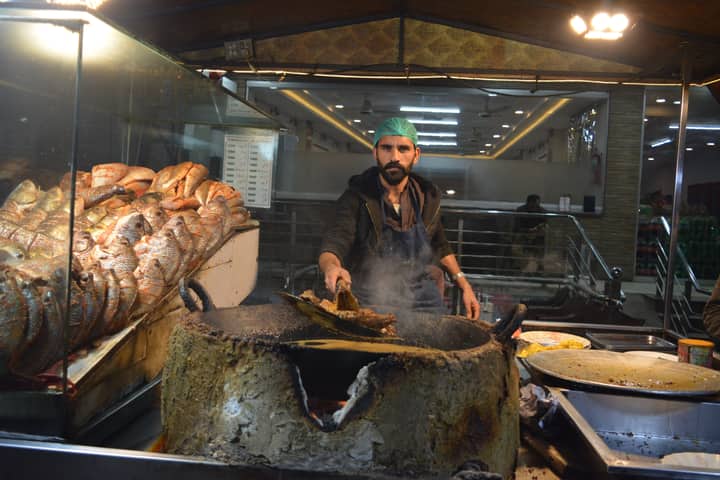 Fish vendor at a Pakistan market frying seafood for customers under bright lights.