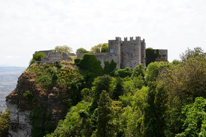 A stunning view of a medieval castle perched on a lush clifftop in Erice, Sicily, surrounded by vibrant green foliage.