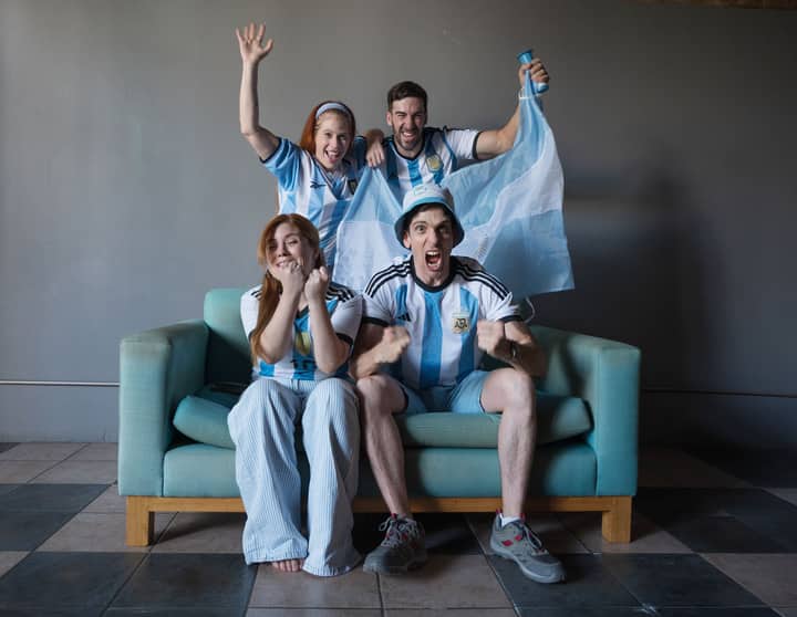 Group of enthusiastic fans in Argentina jerseys watching a soccer match indoors.