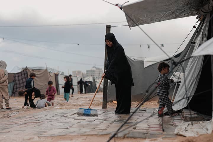 A woman sweeps in a Gaza refugee camp with children playing nearby.