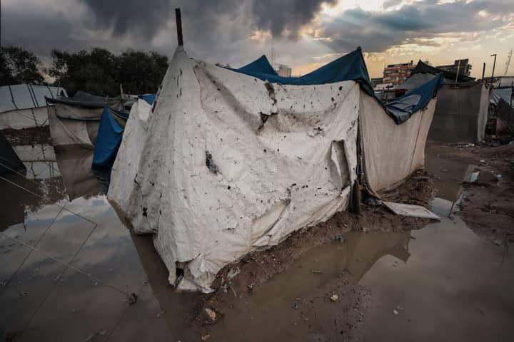 A damaged tent shelter in a flooded area of a Gaza refugee camp under stormy skies.