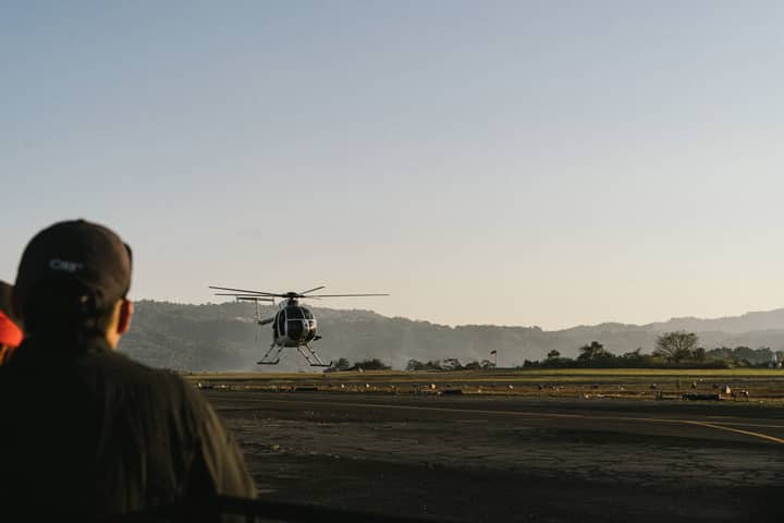 A helicopter takes off at Ilopango Airport in El Salvador, with mountains in the background.