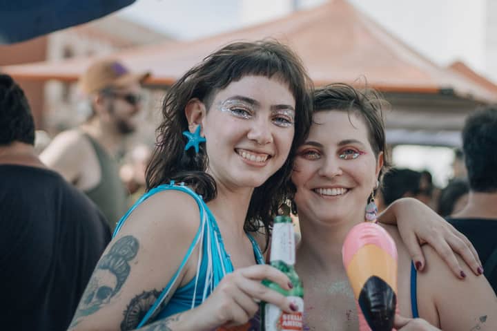 Two friends with glitter makeup joyfully celebrate outdoors, sharing festive drinks.