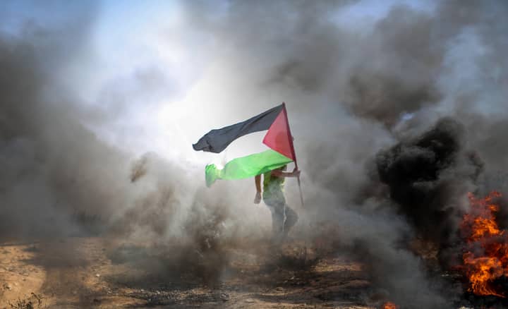 A protestor carrying a Palestinian flag amidst smoke and flames in Gaza.