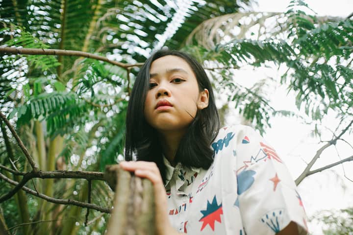 A young woman in a printed shirt surrounded by tropical greenery, looking confidently at the camera.