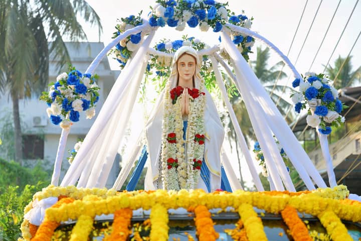 Statue of Our Lady of Lourdes adorned with colorful flowers in an outdoor setting with palm trees.