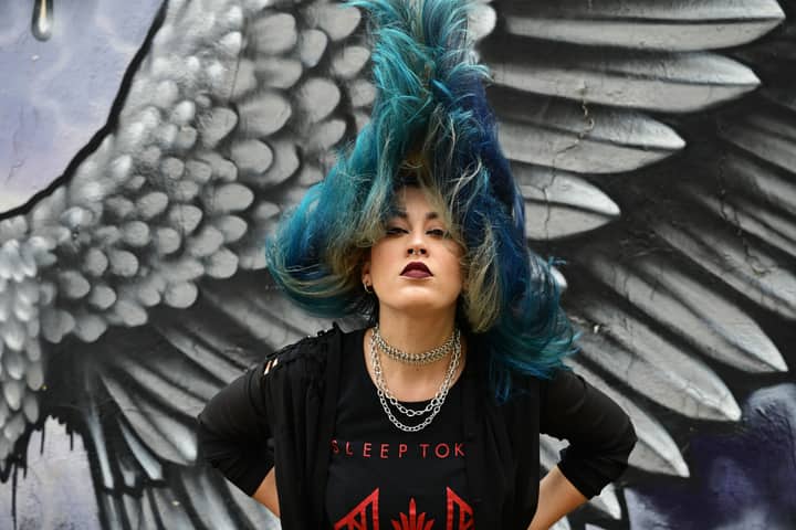 A vibrant portrait of a punk woman with blue hair standing in front of a winged graffiti mural.