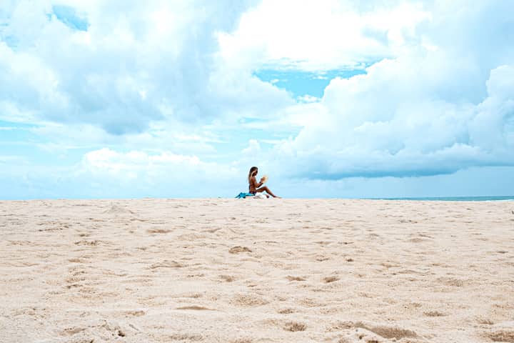 A woman reading a book on the sandy Ponta Negra Beach under a bright sky.