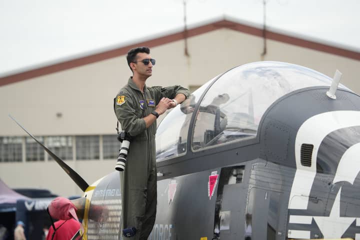 A military pilot in uniform stands confidently atop a modern fighter jet at an airshow.