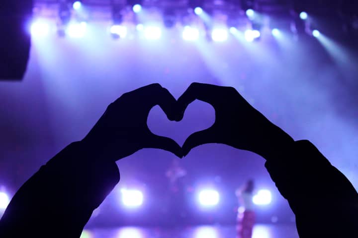 Silhouette of hands forming a heart against bright stage lights at a concert.