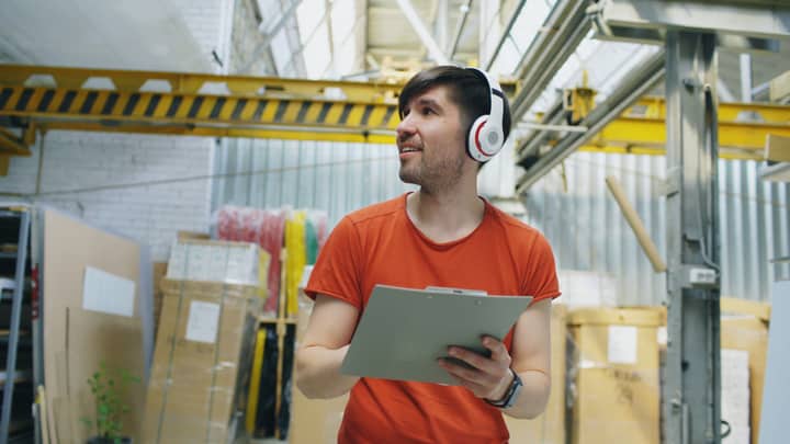 Young man in warehouse wearing red shirt and headphones, holding clipboard.