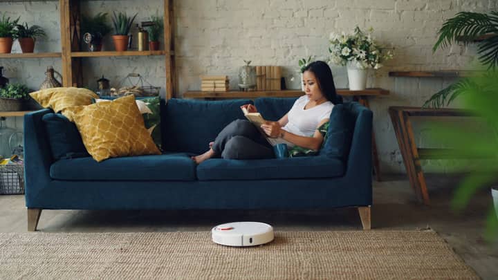 Woman reading on sofa while a robot vacuum cleans the floor in a cozy living room.