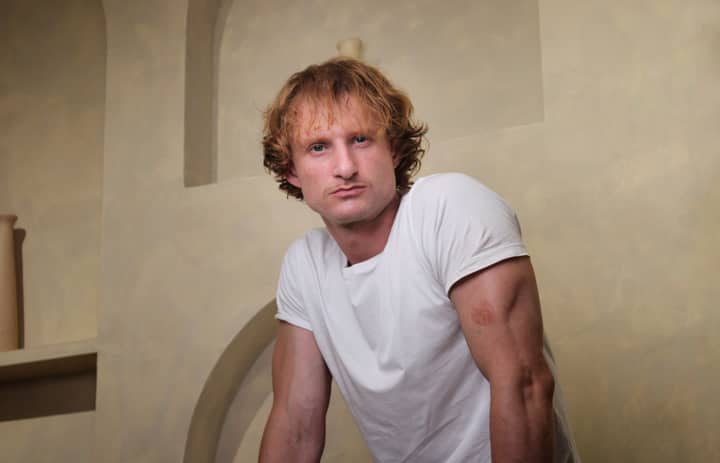A contemplative man in a white t-shirt leans against a wall indoors.