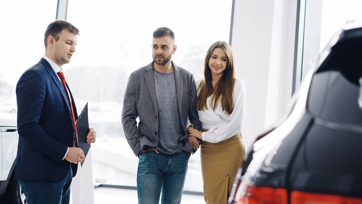 Smiling couple with salesman in car showroom discussing features of a car.