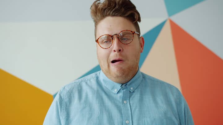 A young man with glasses makes a humorous expression against a colorful geometric wall.