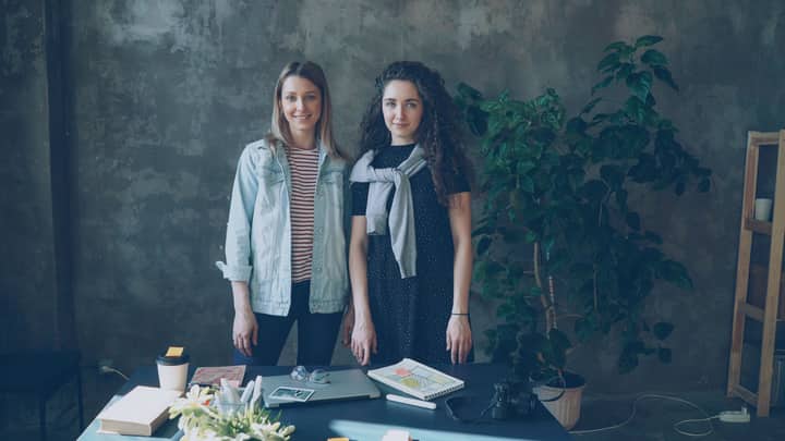 Two women in casual attire standing indoors by a desk, creating a cozy workspace vibe.