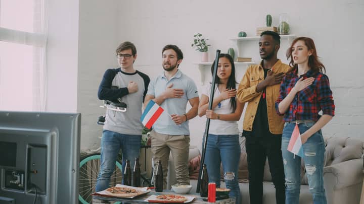 Five friends watching sports, hand on heart, holding flags, showing support indoors.