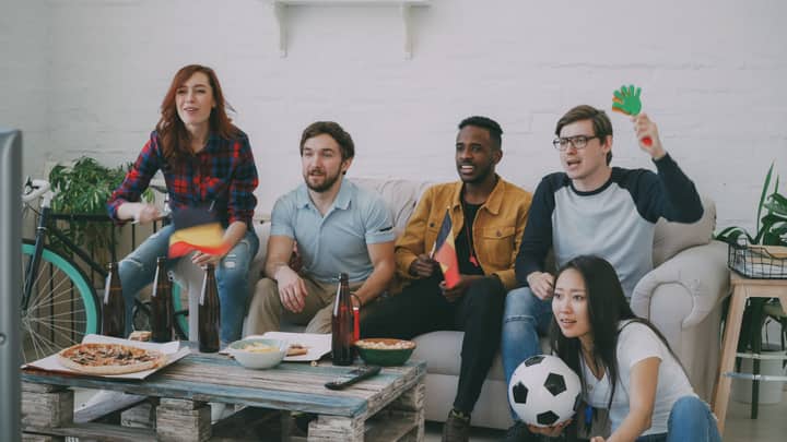 Friends watching a soccer match indoors, waving flags and cheering with snacks and drinks.