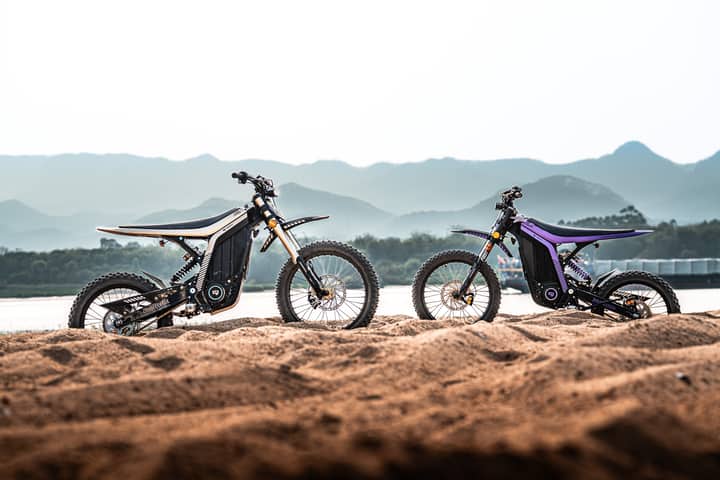 Two electric dirt bikes parked on sandy terrain against a scenic mountain backdrop during sunrise.