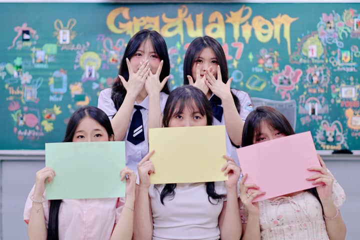 Five students hold colorful papers in front of a decorated blackboard during a graduation celebration.