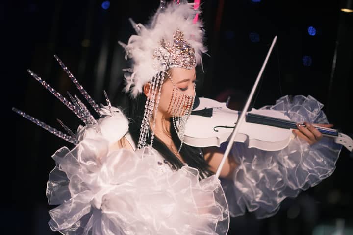 An artistic performer in elaborate costume plays a white violin under stage lights, creating a vibrant spectacle.