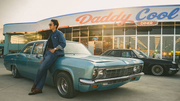 Man in denim outfit leans on vintage car at 'Daddy Cool' diner in retro setting.