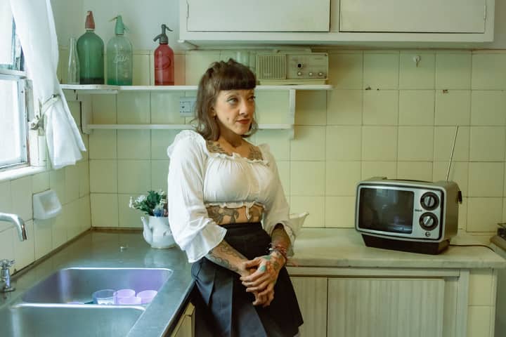 Tattooed woman in a vintage kitchen with retro decor and appliances, exuding a classic vibe.