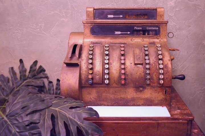 A vintage cash register with a rustic, aged appearance alongside tropical foliage.