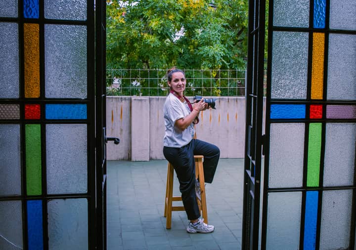 A woman photographer sitting on a stool holding a camera on a balcony framed by a stained glass door.