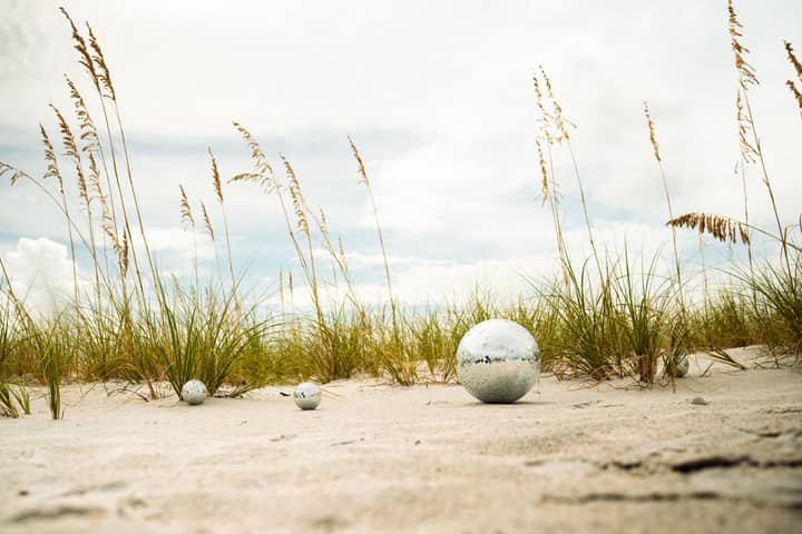 Serene beach with ornamental reflective spheres and tall grass under a cloudy sky.