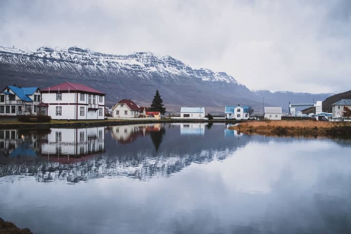 Picturesque Icelandic village with reflections on a calm lake, against a mountainous backdrop.