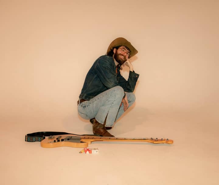 A stylish man in denim and cowboy hat crouches beside an electric guitar in a neutral-toned studio.