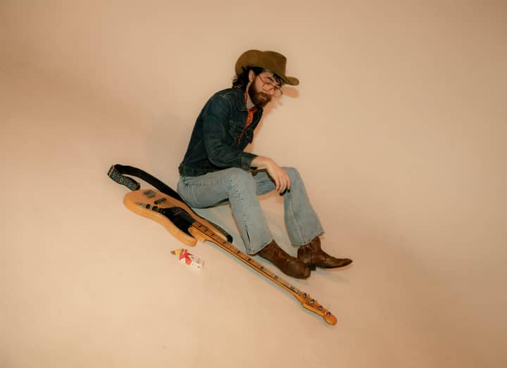 Young male musician in cowboy hat and denim posing with electric guitar in a studio setting.