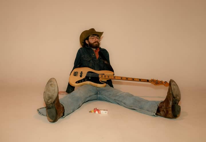 A relaxed musician with a bass guitar, sitting casually with legs apart, wearing jeans and a cowboy hat.