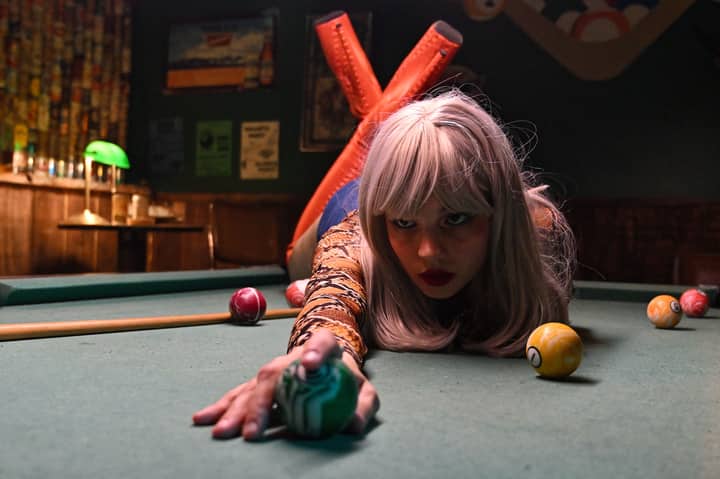 A woman with a striking look poses confidently on a pool table, holding a billiard ball.