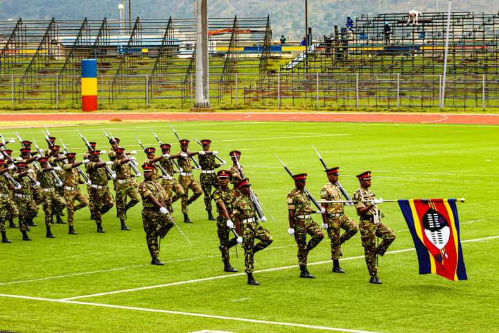 A group of soldiers performing a ceremonial march on a field in Eswatini, holding national flags.